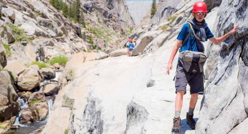 A young person wearing safety gear navigates rocky terrain near a creek. 